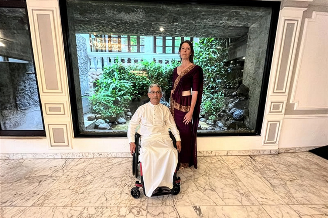 A person in a wheelchair wearing white full body clothing with a person standing wearing a maroon and gold dress