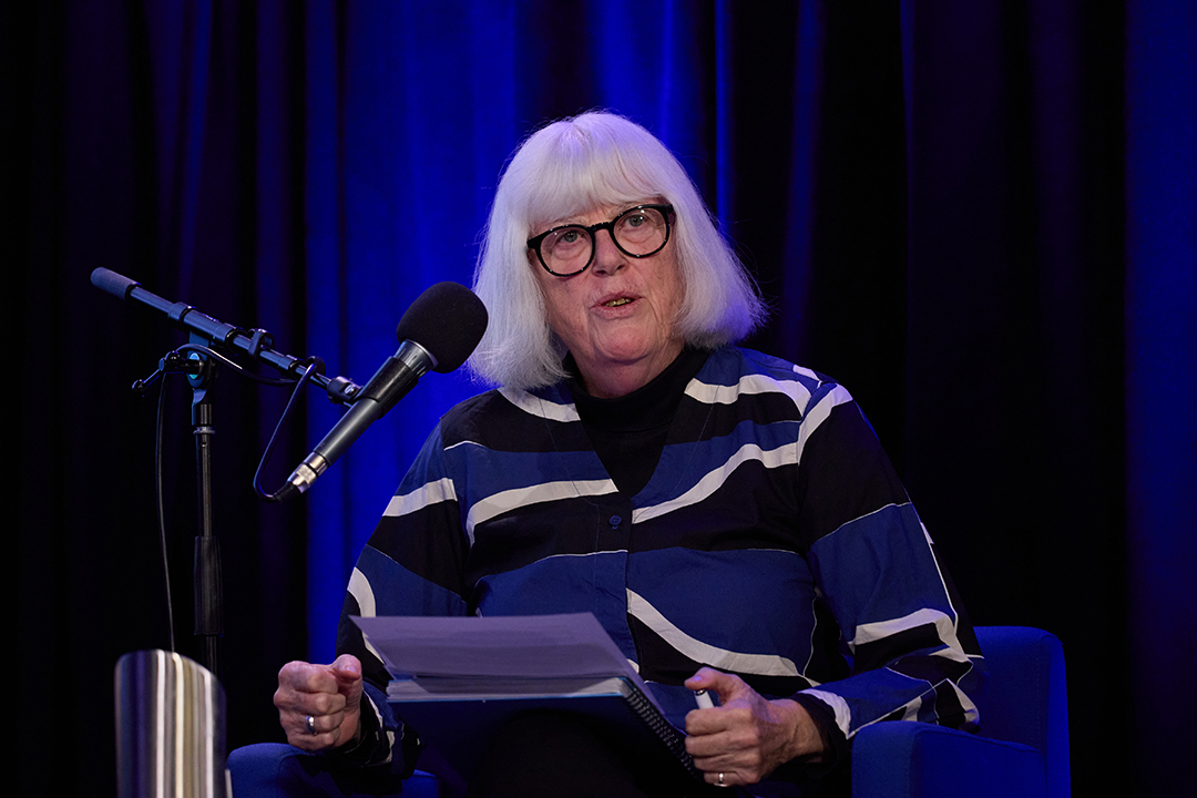 A person with short white hair sitting on a stage in front of a microphone.