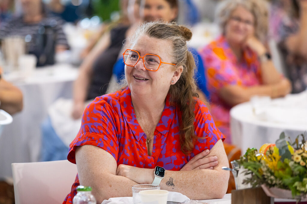A person wearing brightly coloured clothes and glasses sitting at a table in a room full of people