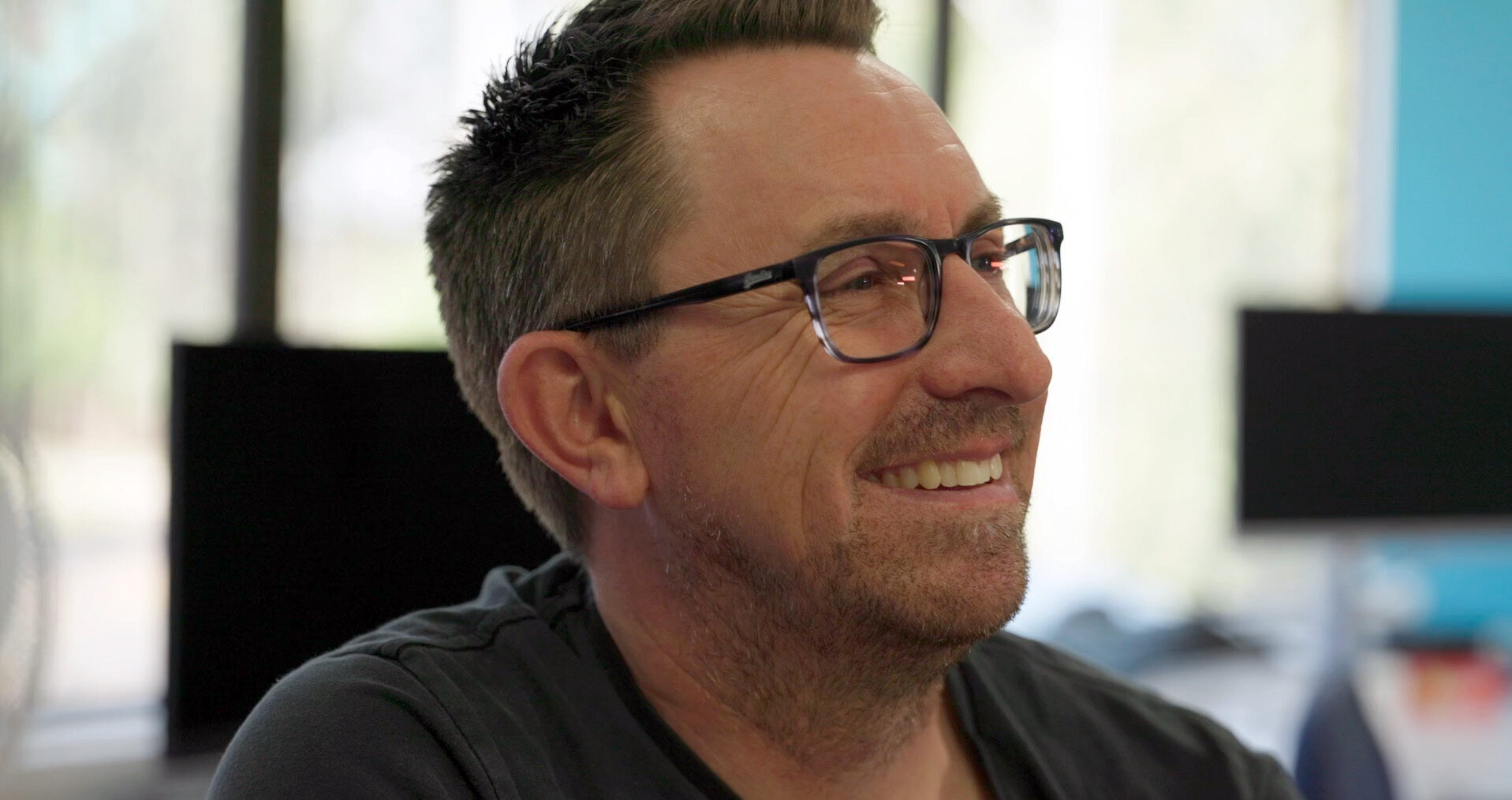 A person wearing dark shirt and dark glasses, smiling in an office setting