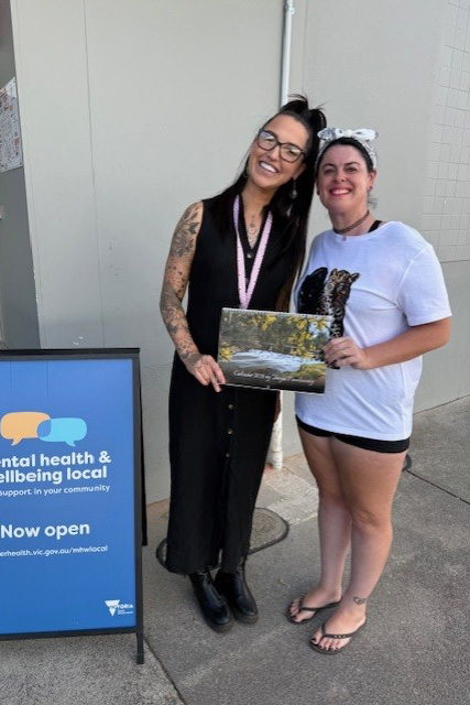 Two people posing for a photo, holding a calendar next to a blue A-frame sign