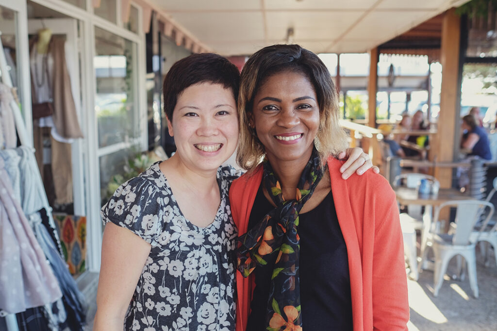Two people at a cafe smiling and hugging. One is wearing a black and white floral shirt and the other is wearing a soft red cardigan.