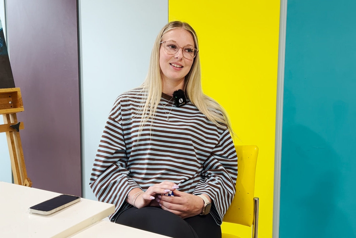 A person with long hair and a striped shirt sitting in front of a blue, yellow and teal wall