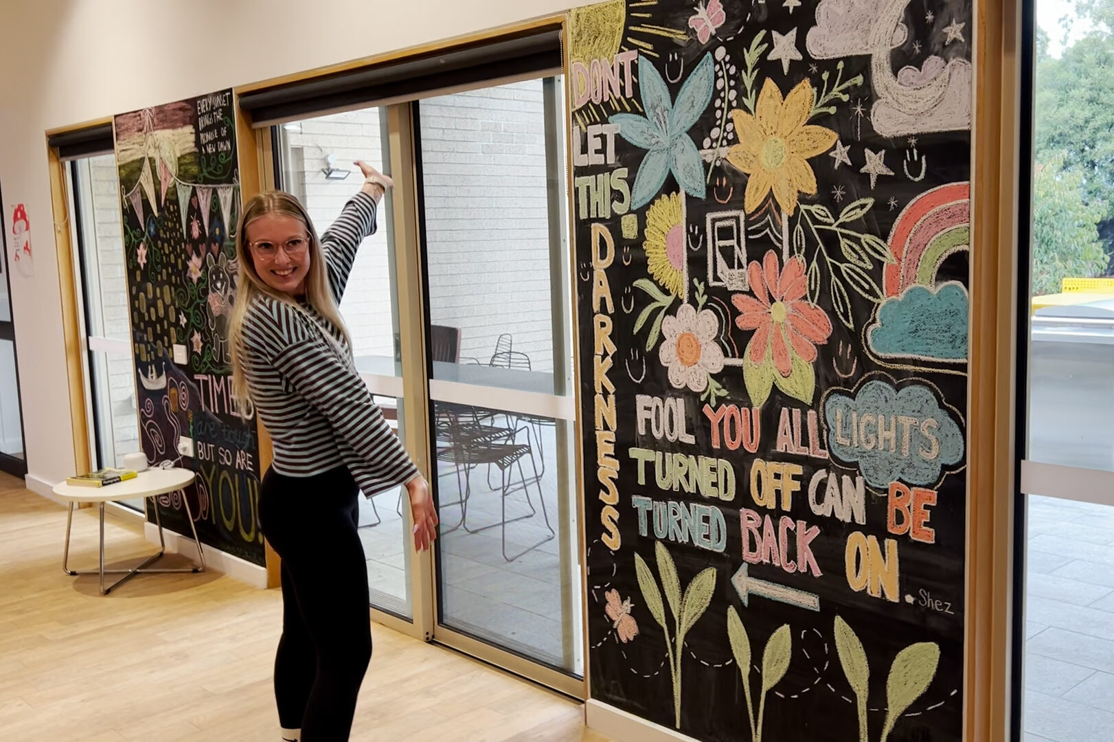 A person smiling and standing next to a colorful wall mural with their arms out