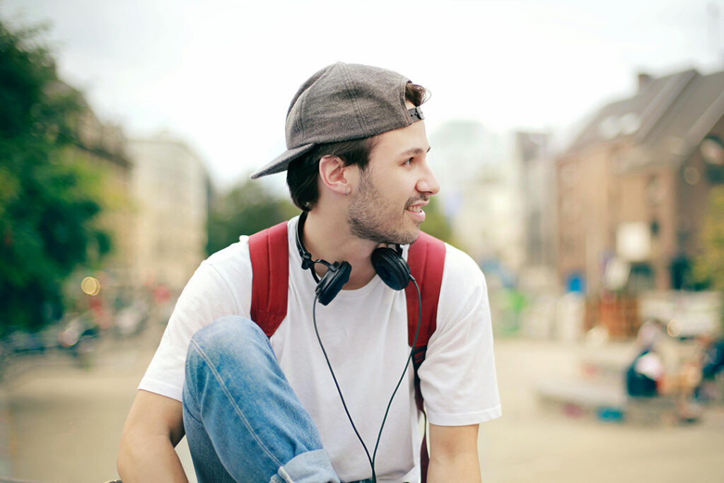 A young person wearing a backwards cap, headphones anda backpack on a busy urban street.