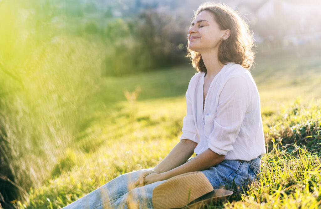A smiling person wearing jeans and a loose shirt sitting in the grass in the sun