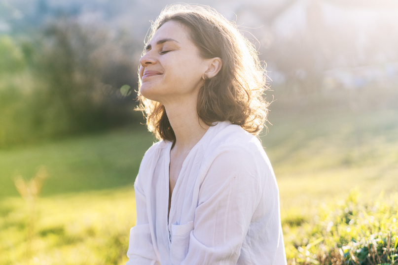 A smiling person wearing jeans and a loose shirt sitting in the grass in the sun