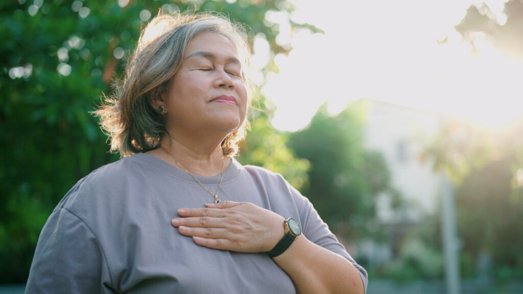 A person with medium length wavy hair meditating in the park with one hand resting on their chest