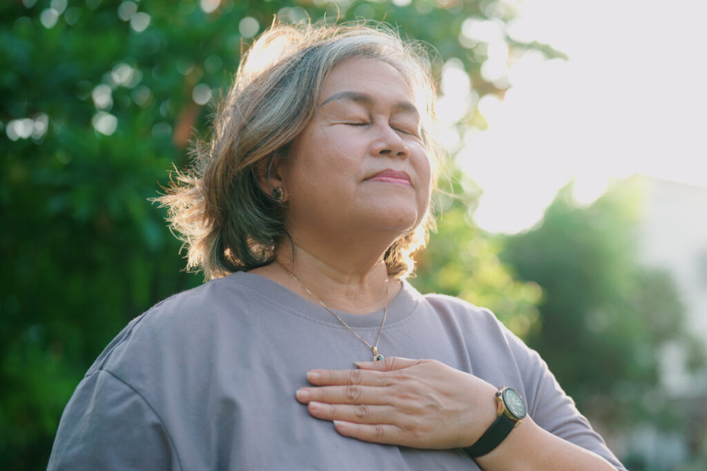 A person with medium length wavy hair meditating in the park with one hand resting on their chest