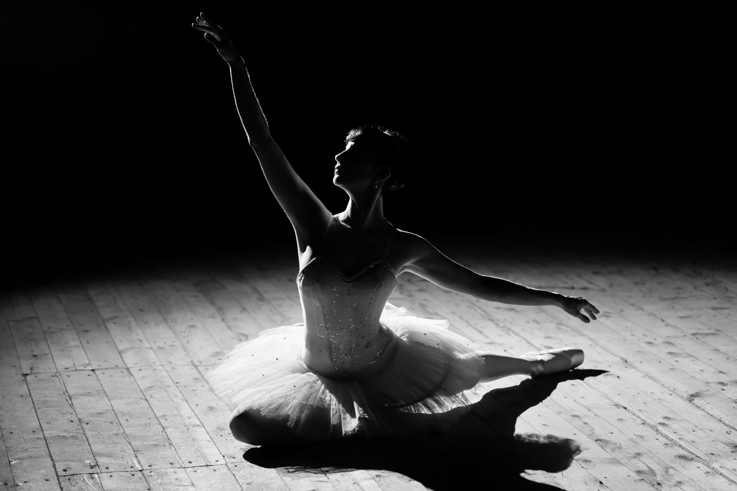 A moody black-and-white photograph of a ballerina seated on a stage floor, posing gracefully under soft dramatic lighting.