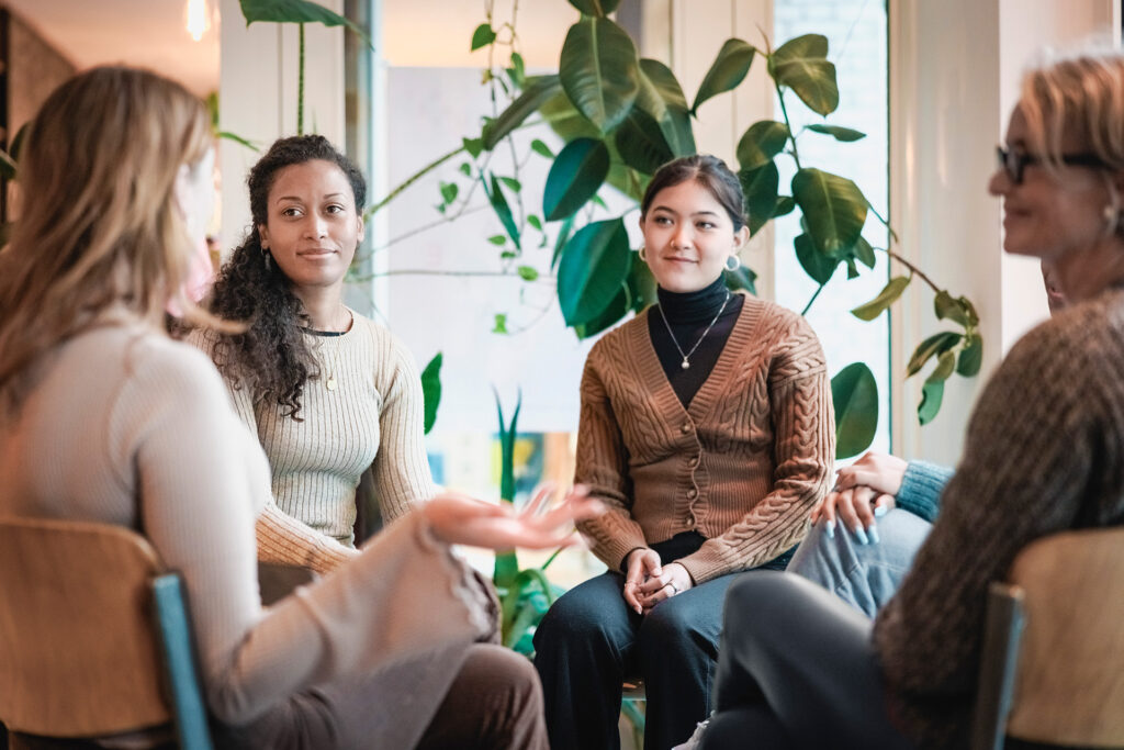 Four people sitting in a circle facing one another in a modern office setting with a large indoor plant in the background.