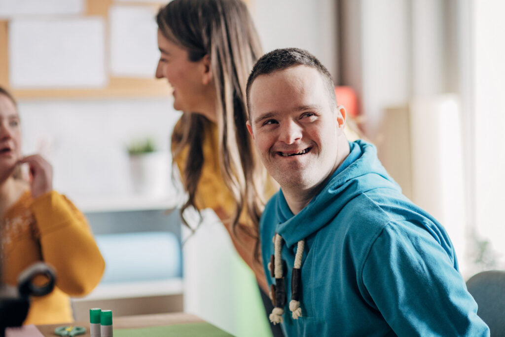 A young person wearing a hooded jumper smiling while other people in the background have a conversation.
