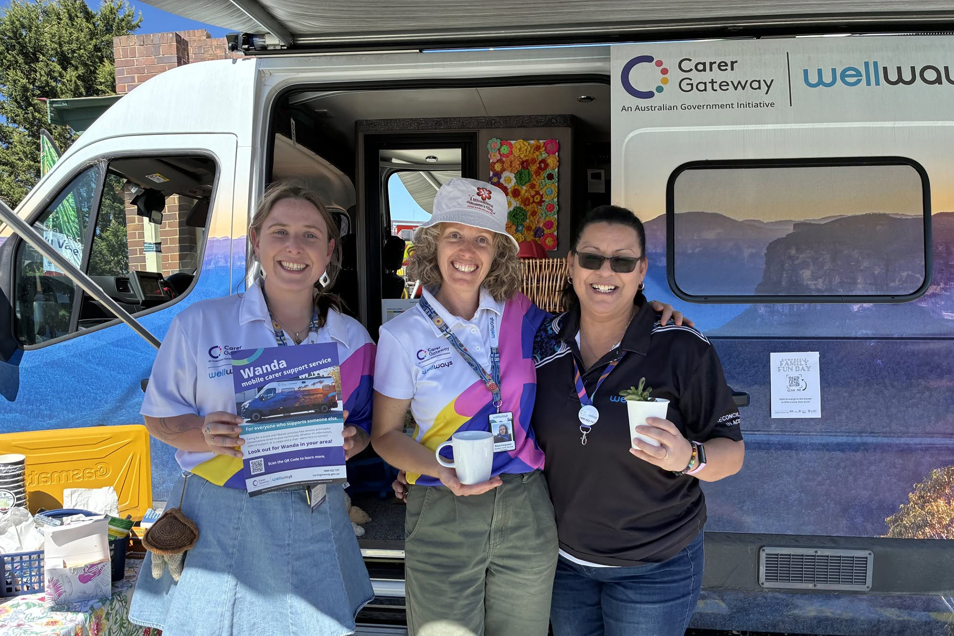 Three people, wearing branded attire, standing under the awning of a large colourful van with a Wellways Carer Gateway logo and an image of mountains covering the side panels