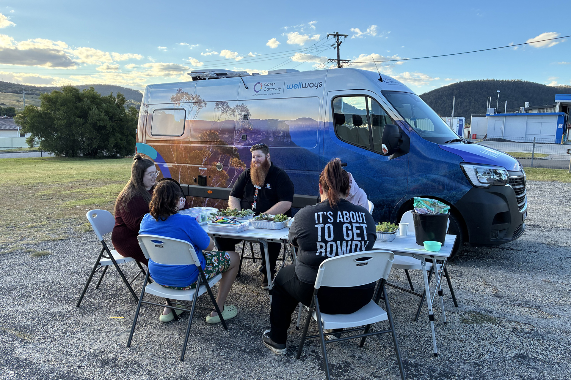 A group of people sitting at fold out tables, next to a large colourful van with a Wellways Carer Gateway logo and an image of mountains covering the side panels