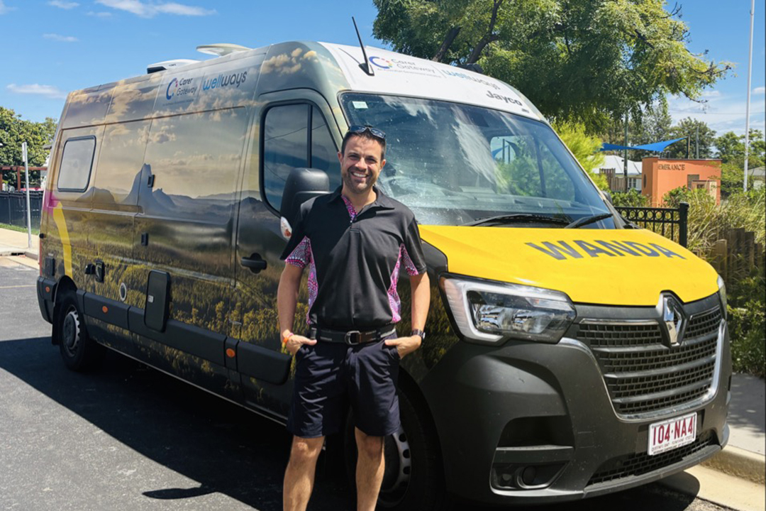 A person wearing a polo shirt and shorts, standing in front of a large colourful van with a Wellways Carer Gateway logo and an image of mountains covering the side panels and the word wanda on the bonnet.