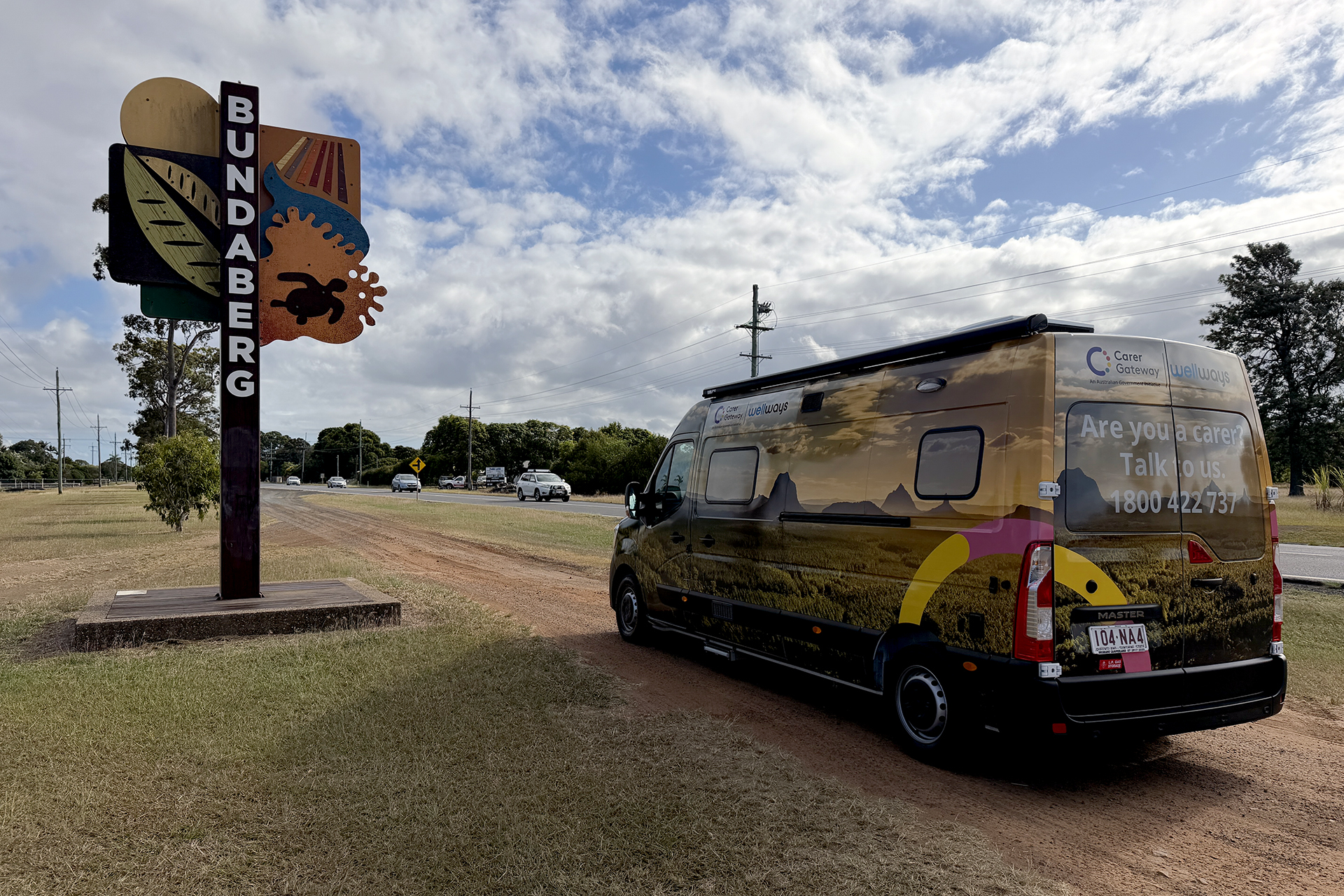 A large colourful van with a Wellways Carer Gateway logo and an image of mountains covering the side panels parked in front of a signpost with colourful artwork and the word Bundaberg,