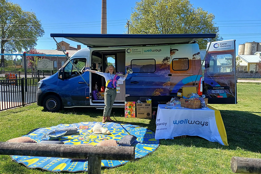 A person with open arms standing under the awning of A large colourful van with a Wellways Carer Gateway logo and an image of mountains covering the side panels