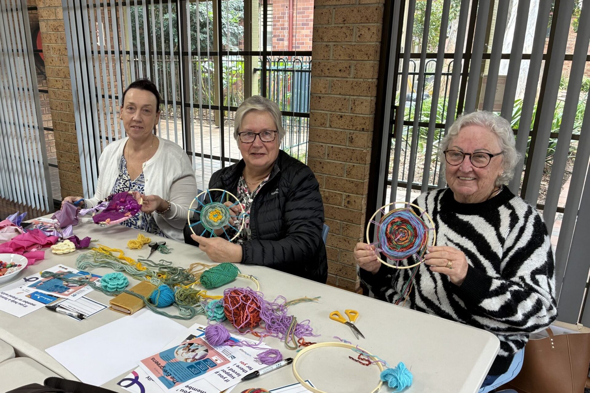 Three people sitting in front of a table with materials on them holding embroidery hoops
