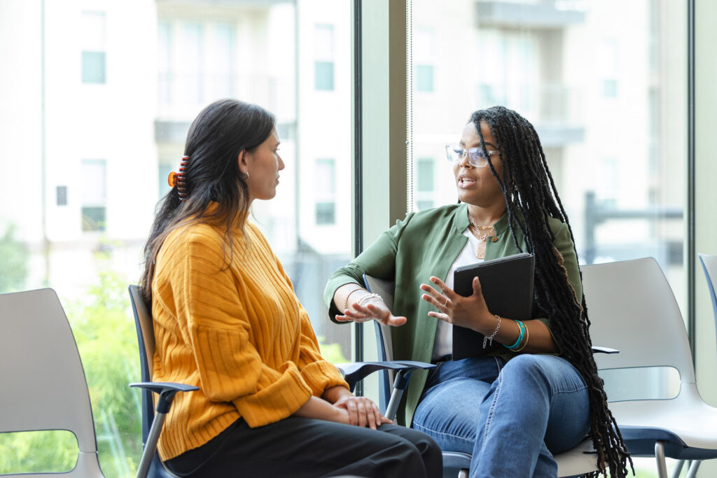Two people sitting infront of a large window in a counseling session. The therapist gestures while giving advice.
