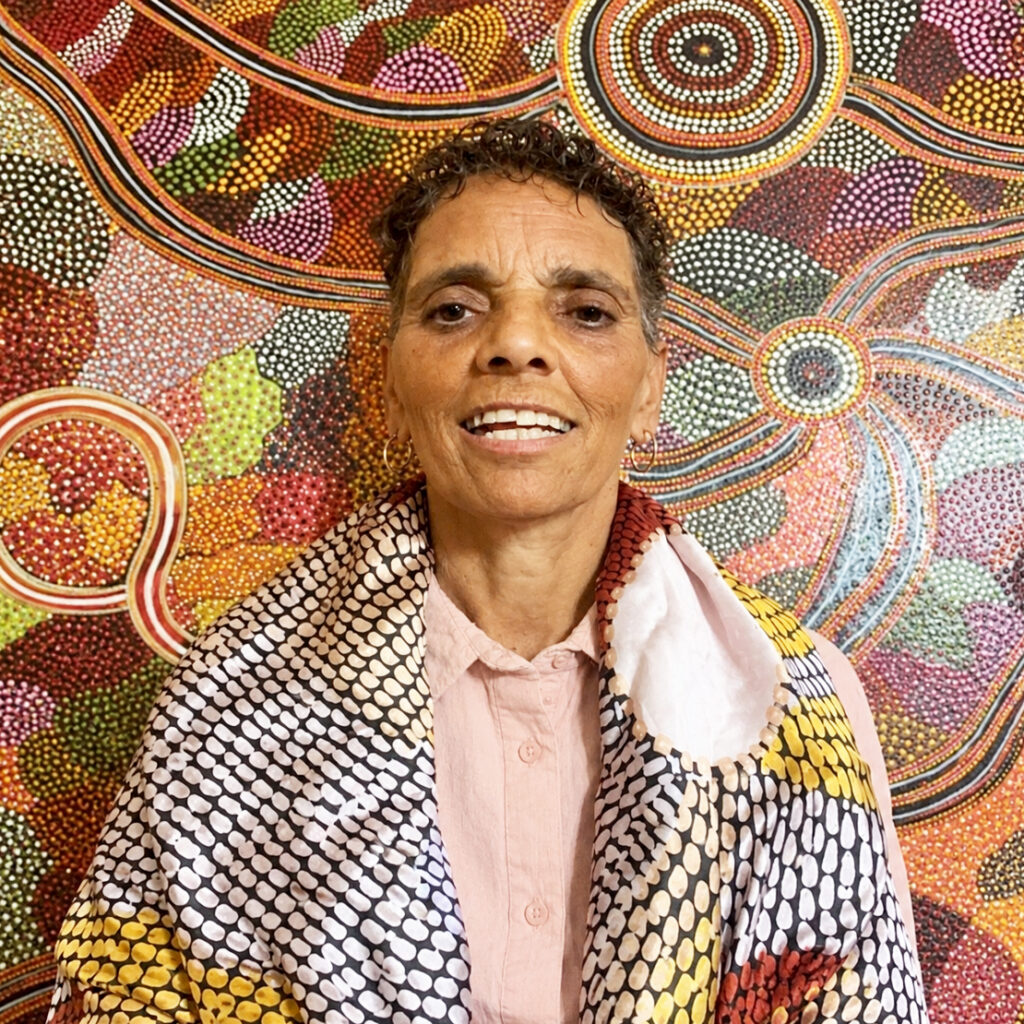 A portrait of a person with short dark hair, wearing a scarf standing in front of a First Nations wall painting